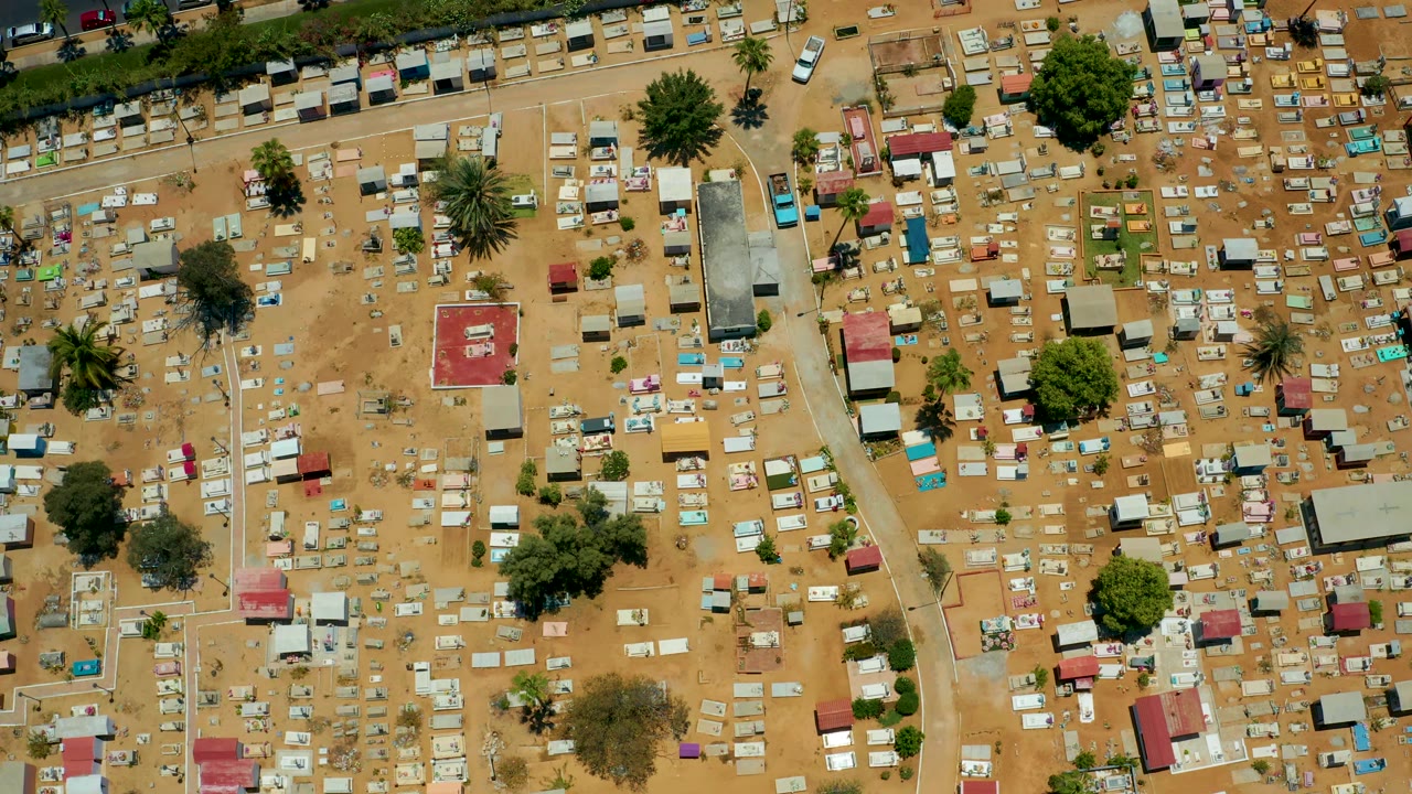 Overhead San Jose Del Cabo cemetery view with red-roofed structures, compact grave markers, sandy lanes, green trees, and no visible people.