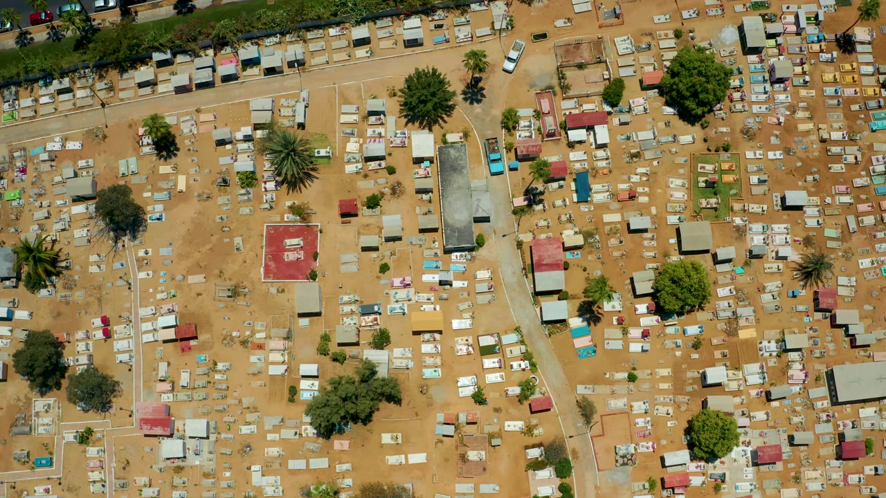 Aerial frame of the San Jose Del Cabo cemetery showing rows of graves, white and colored markers, green trees, a top-edge road, and no visible people.