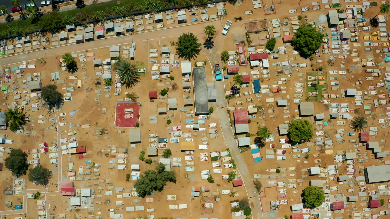 Top-down view over the San Jose Del Cabo cemetery with sandy paths, many tombstones, a few larger trees, roadside vehicles, and no visible people.
