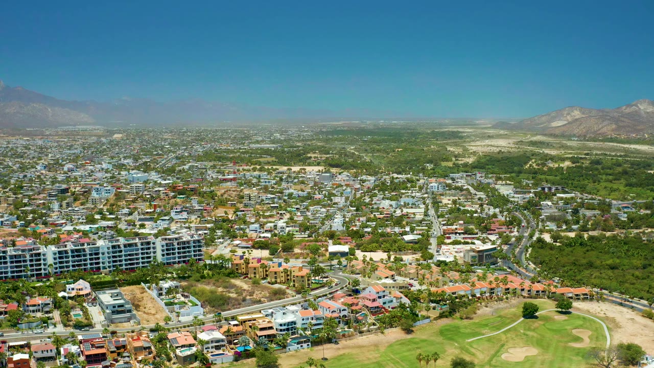 Opening aerial frame over San Jose Del Cabo with a golf course at the bottom edge, residential buildings and roads across the middle, distant mountains, clear sky, and no visible people at this scale.