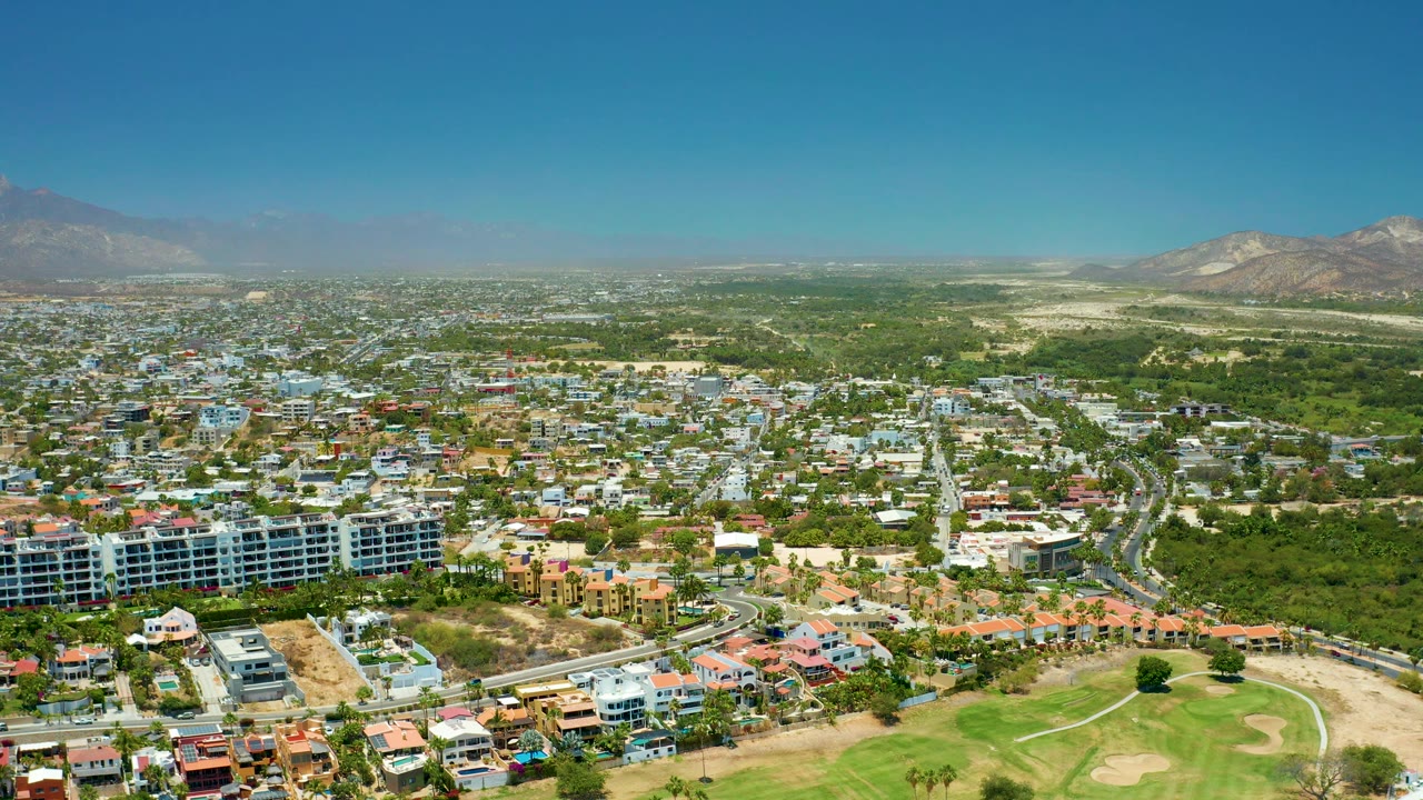 Mid-clip aerial over San Jose Del Cabo showing dense residential streets, a broad green tree belt, pale dry ground, the golf course below, distant mountains, and no visible people at this scale.