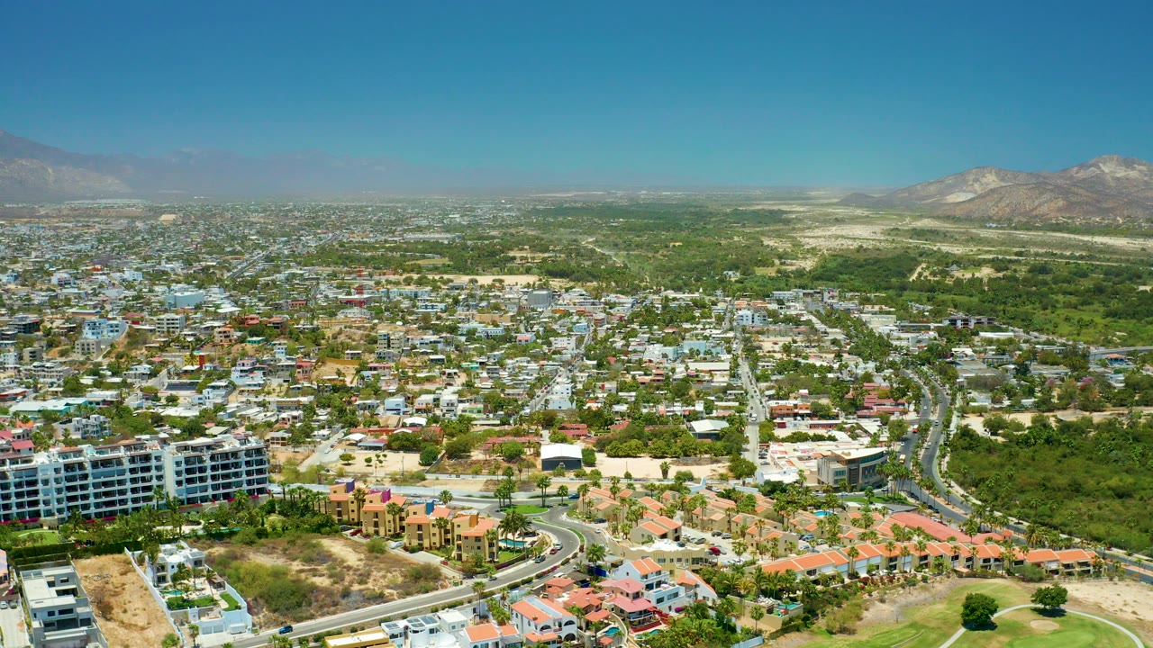 Opening aerial view over San Jose Del Cabo, Mexico, with low-rise buildings, a curved road, green areas, distant mountains, clear blue sky, and no visible people.