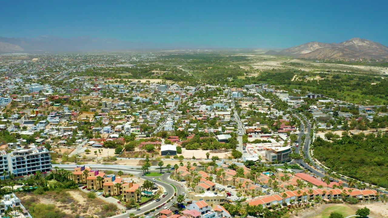 San Jose Del Cabo aerial view at the opening moment, with foreground homes, a curved road, dense greenery, distant mountains, and no visible people.
