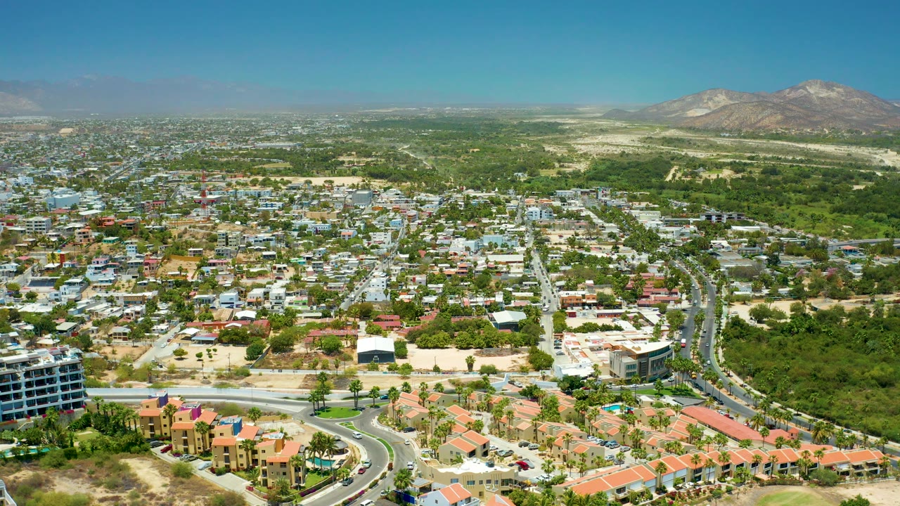 Aerial view across houses, roads, greenery, and distant mountains in bright daylight.