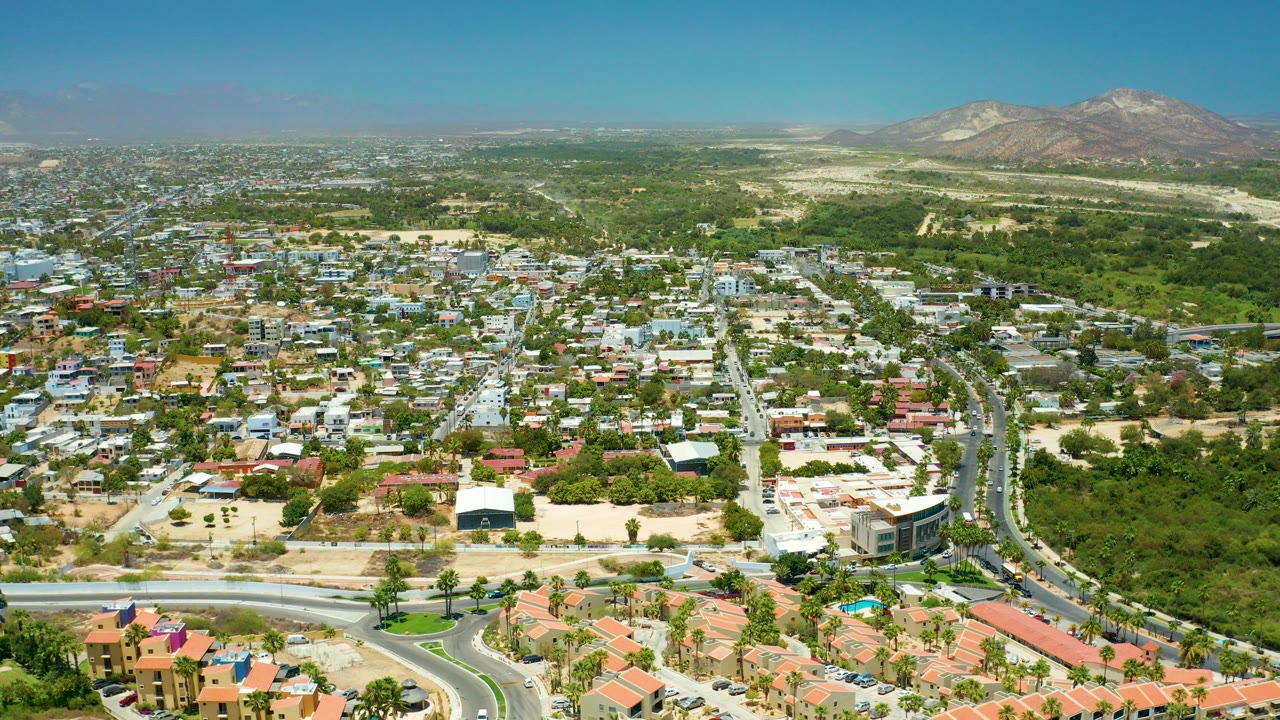 A wide aerial frame of San Jose del Cabo shows houses, curving roads, green open areas, distant hills, bright midday light, and no visible people.