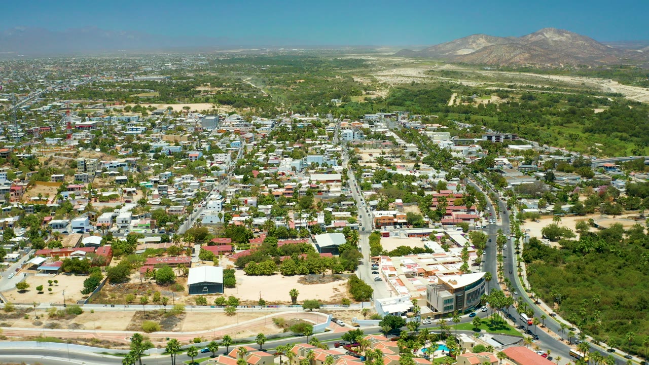Opening aerial frame over San Jose del Cabo houses and streets, with a green corridor through the center, mountains on the horizon, clear blue sky, and no visible people.