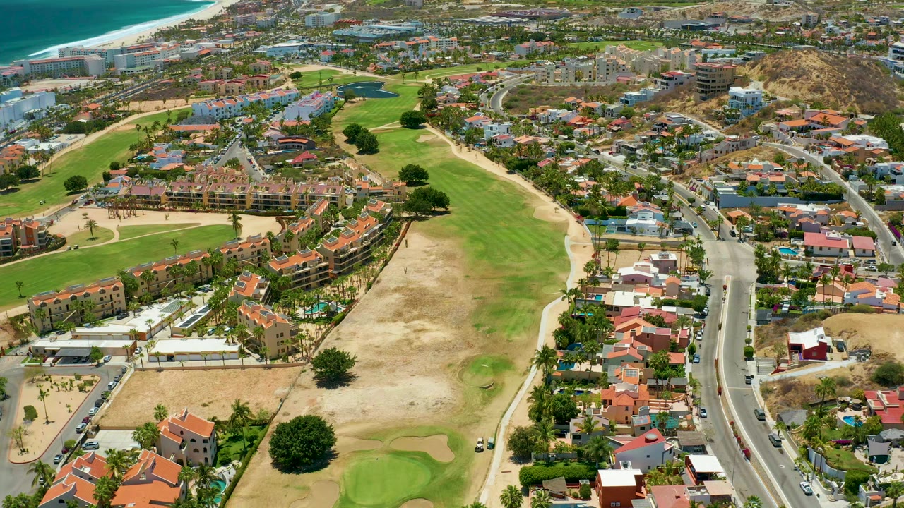 Opening aerial frame over San Jose Del Cabo showing green golf fairways, sand traps, red-tiled homes, a curving road, dry hillside terrain, and ocean at the upper left with no visible people.