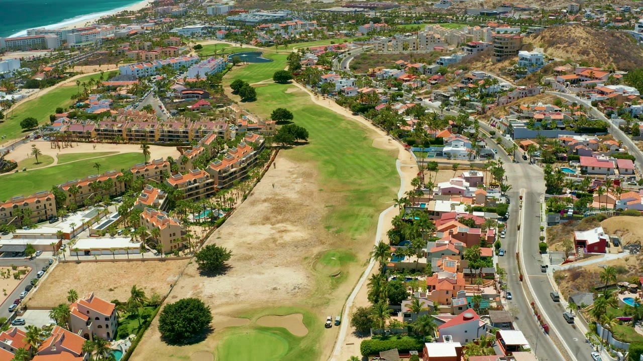 Elevated San Jose del Cabo view with a tan and green golf course on the left, a line of homes and a road on the right, distant coastal development, blue water, and no visible people.