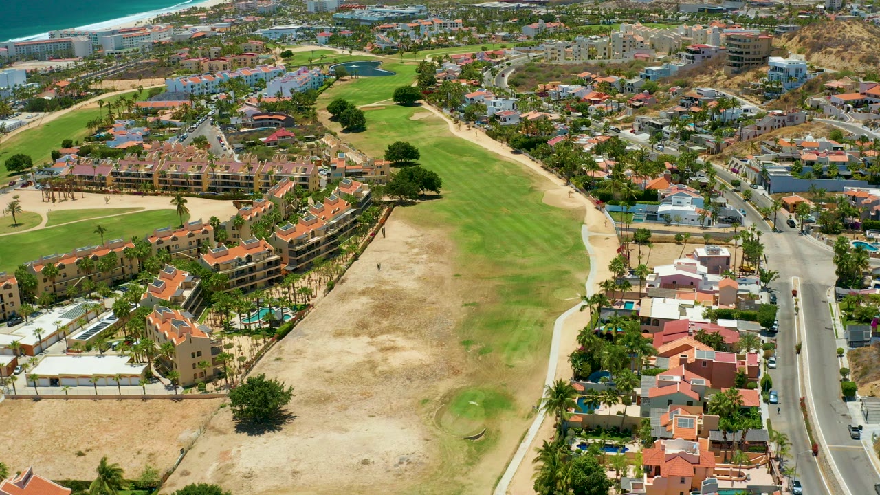 Aerial frame from San Jose del Cabo showing a green and dry golf course bordered by red-roofed residences, a road, distant beach and ocean, and no visible people.