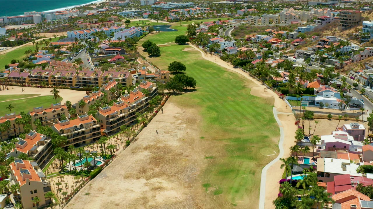 Aerial frame over San Jose Del Cabo showing red-roof residential buildings in the foreground, a green golf fairway, tan sand areas, palms, and blue ocean beyond, with no visible people.