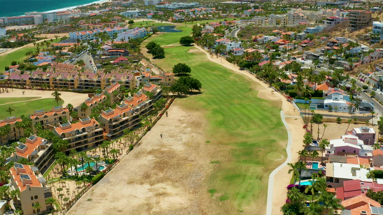 A second aerial view of San Jose Del Cabo keeps the fairway centered while red-roof residences line the lower edge, sandy ground sits left of the grass, and ocean remains visible at the top, with no visible people.