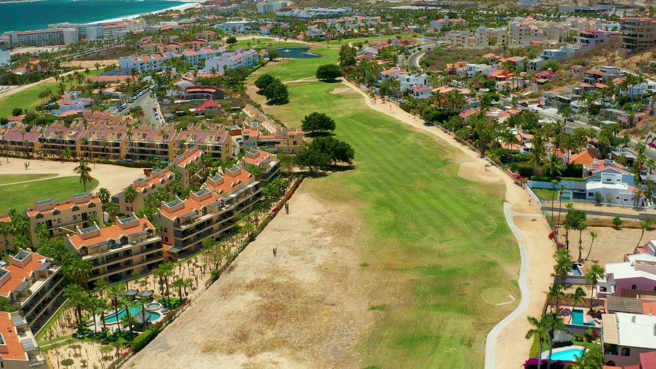 Aerial view across apartments, golf greens, sandy ground, roads, and ocean.