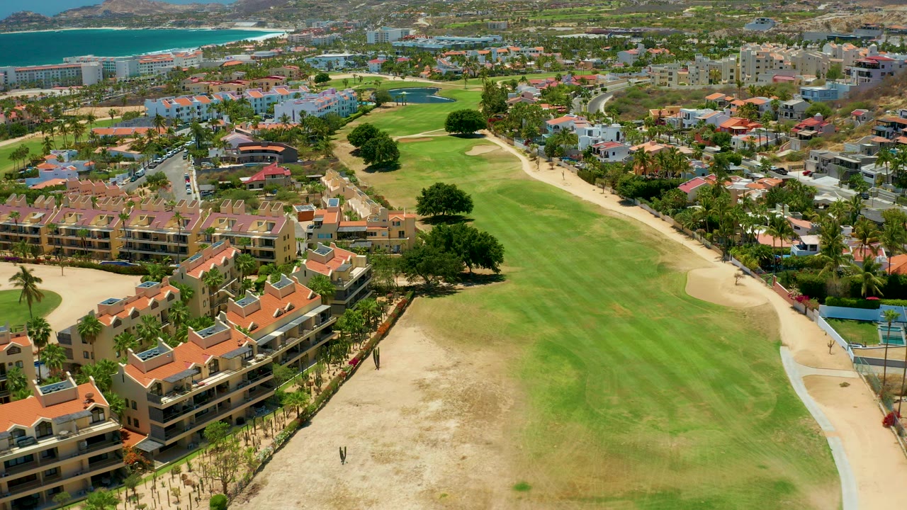 Aerial frame over San Jose del Cabo showing terracotta-roof residences, a green fairway, sand bunkers, distant blue ocean, hills, and a tiny visible person near the lower course edge.