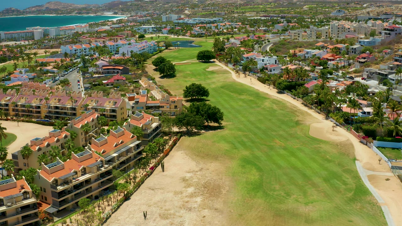 Aerial frame in San Jose del Cabo with the fairway shifting right, sand bunker curves in the foreground, terracotta residential roofs at lower left, blue ocean at upper left, and no clearly identifiable faces.