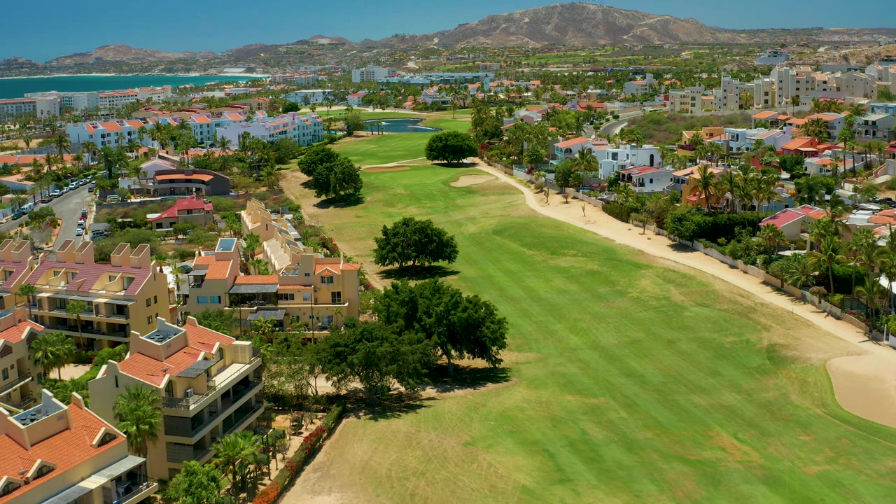 At two seconds, San Jose Del Cabo residential blocks and a wide golf fairway fill the frame, with blue water to the left, sandy bunkers on the course, distant mountains, and no visible people.