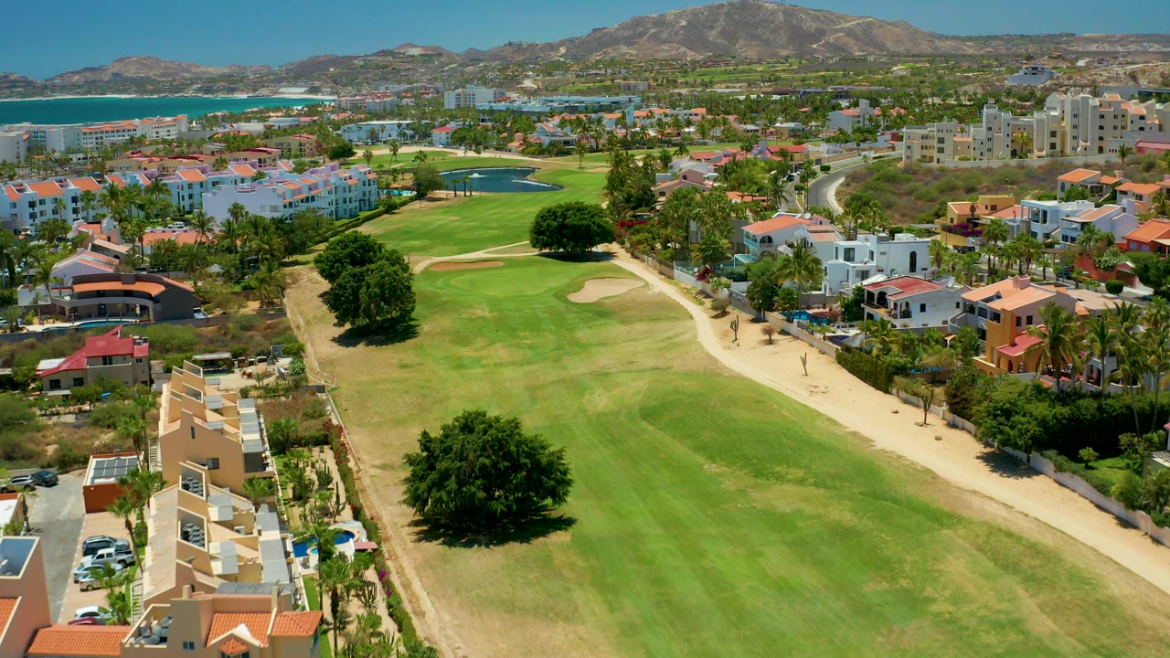 Aerial frame over San Jose Del Cabo showing a green golf fairway, sandy path, red-roofed and white buildings, blue ocean, distant mountains, and no visible people.