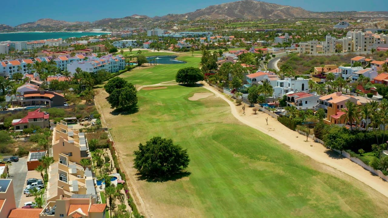 San Jose Del Cabo aerial view with the fairway centered, a large tree near the lower course, residential buildings on both sides, ocean at left, mountains in the distance, and no visible people.