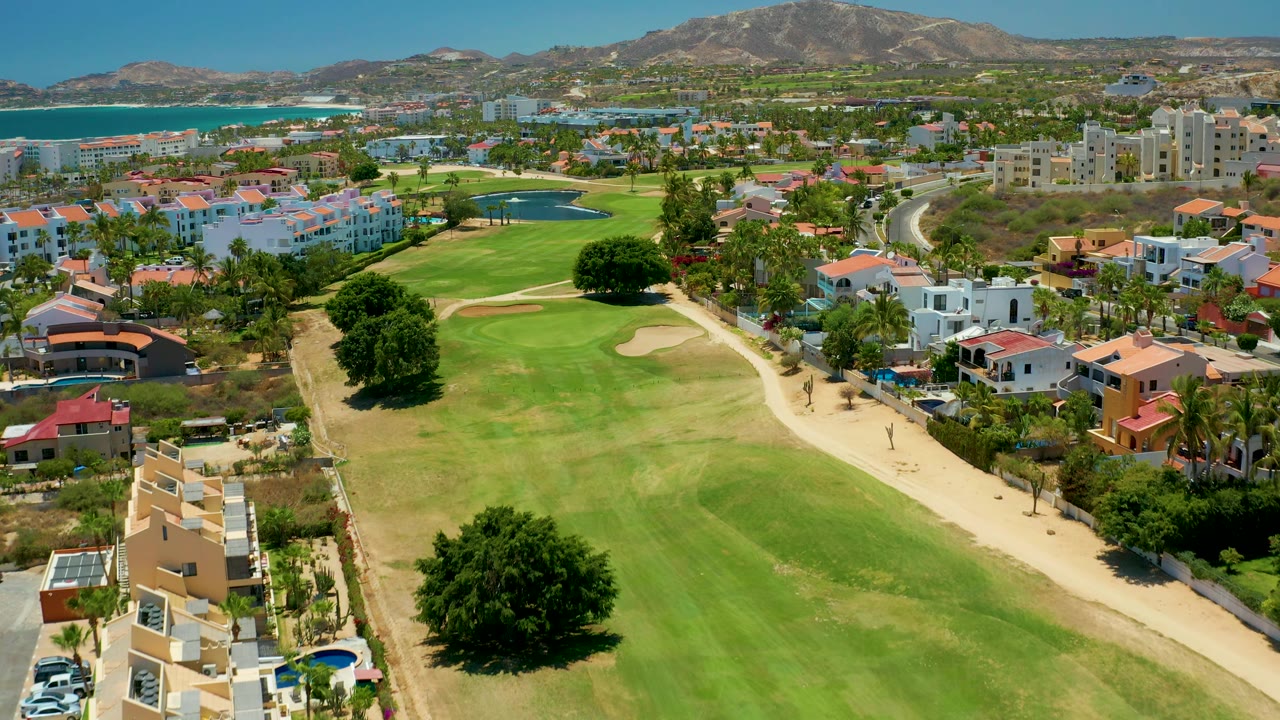 Aerial view in San Jose Del Cabo showing a golf green and sand trap in the foreground, villas and apartment blocks beyond, turquoise ocean at left, mountain ridges behind, and no visible people.