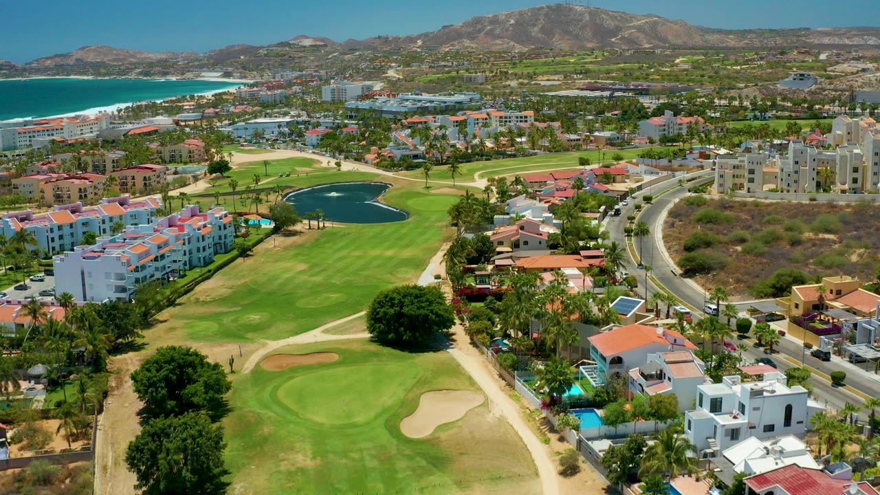 Aerial frame over San Jose del Cabo showing a green golf course with sand bunkers, a pond, coastal residences, blue ocean, sandy beach, and mountains in the distance, with no visible people.