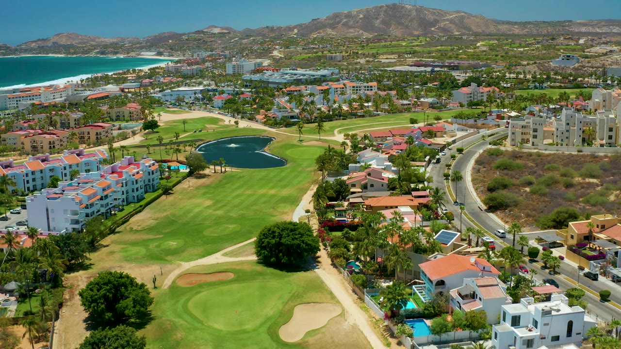 Aerial view in San Jose del Cabo showing the golf course pond slightly left of center, a curving road with small vehicles on the right, clustered residences, the ocean, beach, and mountain ridge, with no visible people.