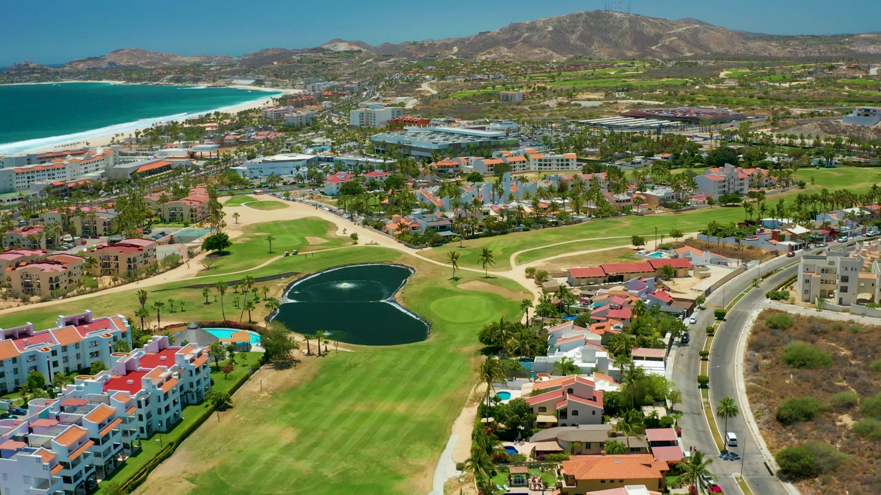 Aerial frame over San Jose del Cabo showing green golf fairways, a dark pond, curved road, red-roofed buildings, beach, ocean, distant mountains, and no visible people.