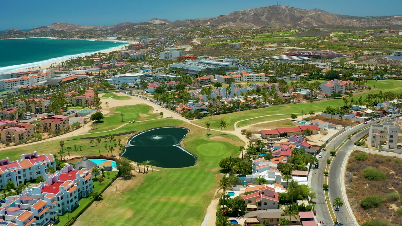 San Jose del Cabo aerial frame showing a golf pond lower right of center, bright fairways, curved road at right, coastal buildings, white beach surf, blue ocean, mountains, and no visible people.