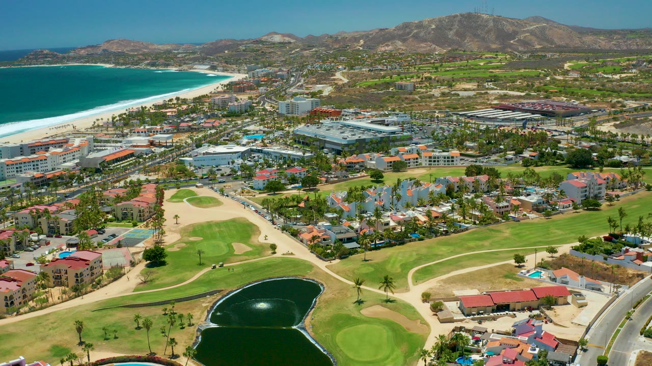 Opening aerial frame over San Jose Del Cabo, Baja California Sur, showing turquoise ocean, white beach, low-rise development, green golf fairways, a dark pond, dry hills, clear sky, and no visible people.