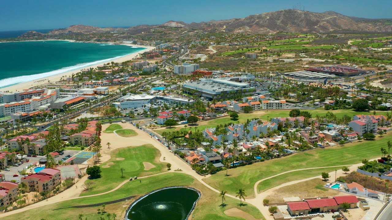 Drone view across beach, roads, buildings, golf greens, pond, and mountain backdrop.