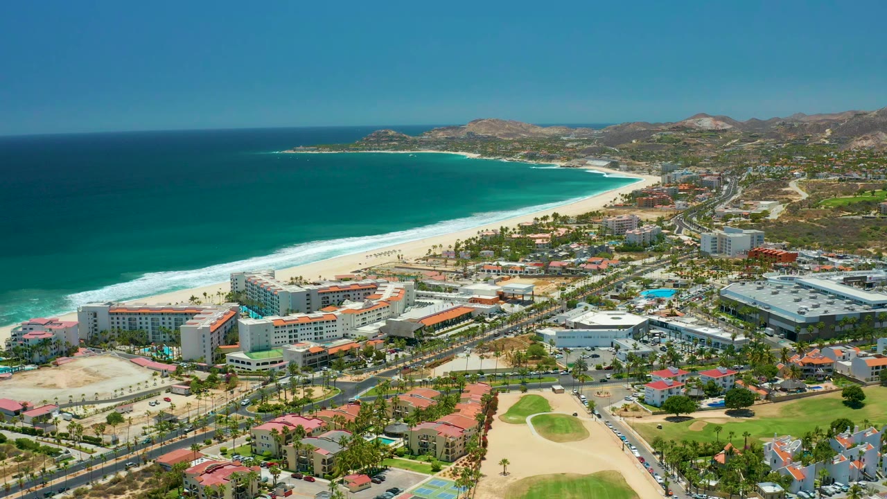 Opening aerial frame over San Jose Del Cabo, Mexico, with turquoise water, white surf, a curving sandy beach, coastal buildings, golf course greens, dry hills, and no visible people.