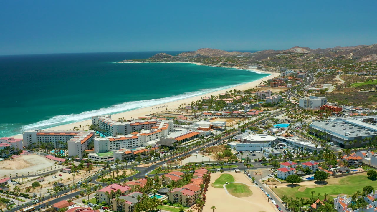 Aerial frame over San Jose del Cabo showing turquoise ocean, a white-sand beach, resort buildings, a coastal road, golf course greens, dry hills, clear sky, and no visible people.