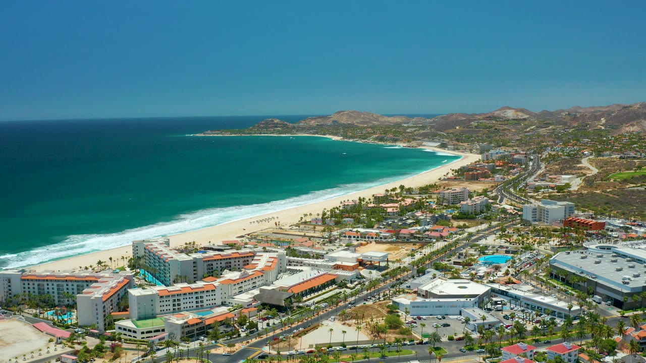 Aerial view of turquoise ocean, sandy beach, resort buildings, roads, pools, and dry hills.