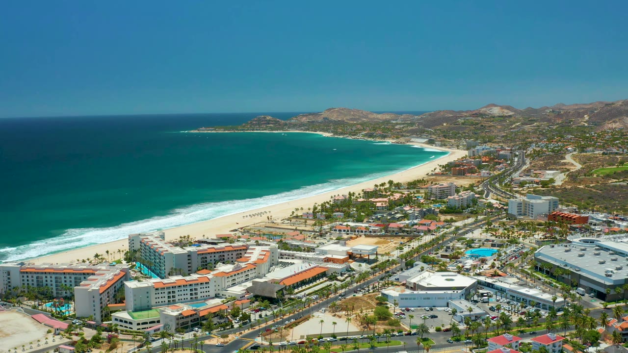 Aerial frame of San Jose Del Cabo, Mexico showing the crescent beach and blue ocean on the left, resort blocks and roads below, pool areas near the buildings, and no visible people.