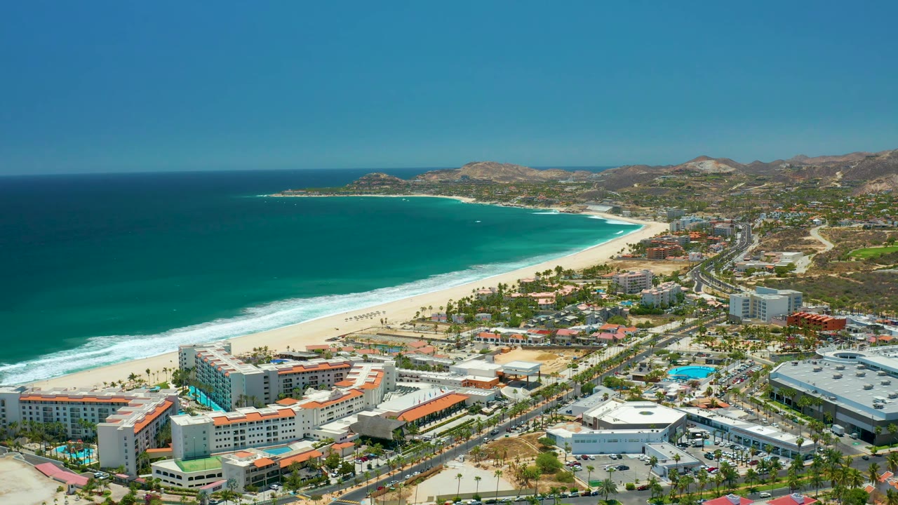 Wide drone-like view in San Jose Del Cabo, Mexico with the beach sweeping across the frame, green-blue ocean offshore, resort roofs and pools below, inland roads and hills to the right, and no visible people.
