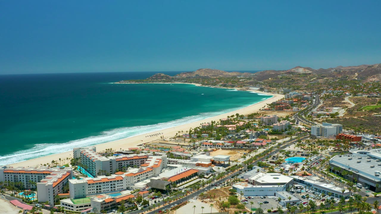 High oblique aerial over San Jose Del Cabo, Mexico with the ocean filling the left side, a bright sandy shoreline, resort buildings and palm rows below, inland roads and hills at right, and no visible people.