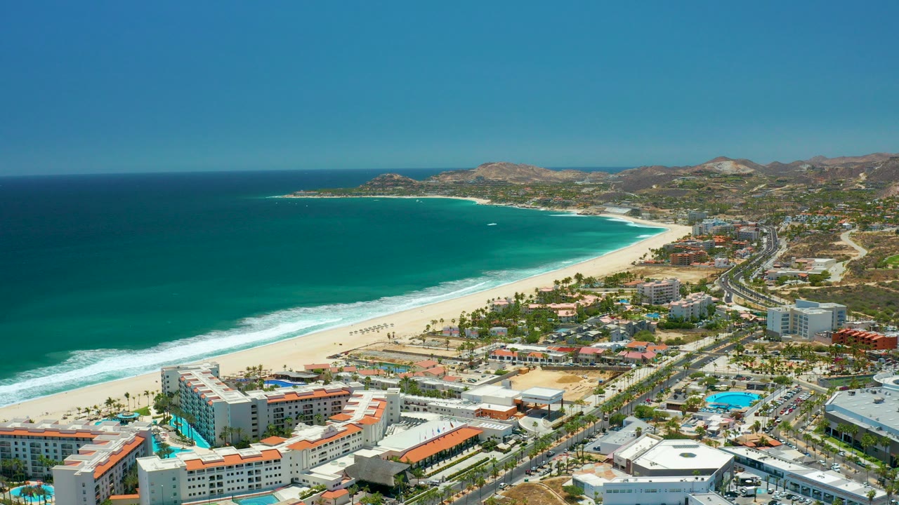 Opening aerial frame over San Jose del Cabo with turquoise ocean at left, white surf on the beach, beachside buildings and road traffic below, dry hills in the distance, and no visible people.