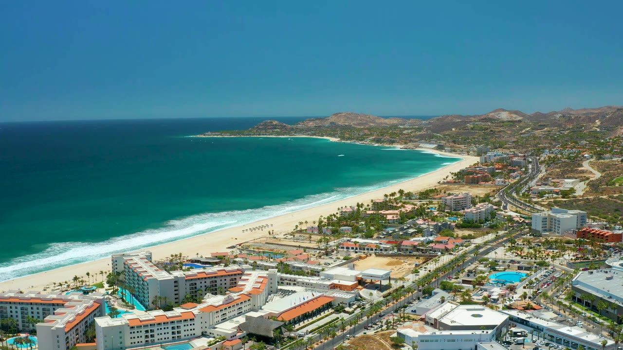 One second into the clip, the San Jose del Cabo beach curves across the frame with the ocean on the left, a parallel road with small vehicles on the right, coastal buildings below, and no visible people.