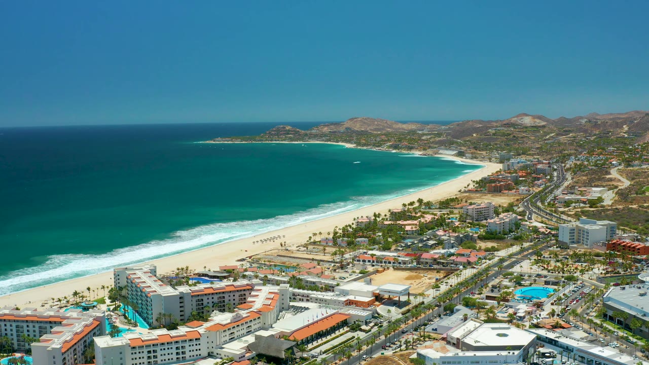 At 0:02, the aerial view holds over San Jose del Cabo with beachfront hotels in the lower frame, surf lines along the sand, highway traffic inland from the beach, hills beyond, and no visible people.