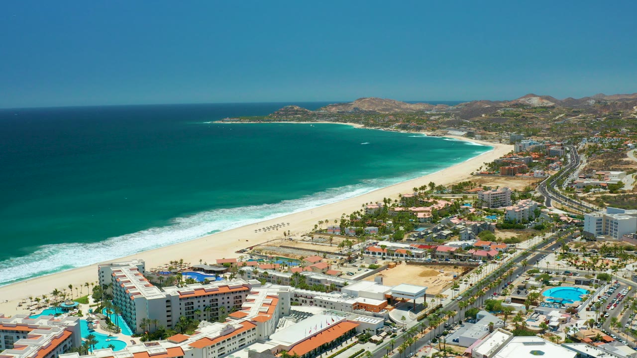 Aerial frame over San Jose Del Cabo with turquoise ocean at left, white surf on the sand, beachfront hotels and pools in the foreground, a coastal road at right, distant hills, and no visible people.