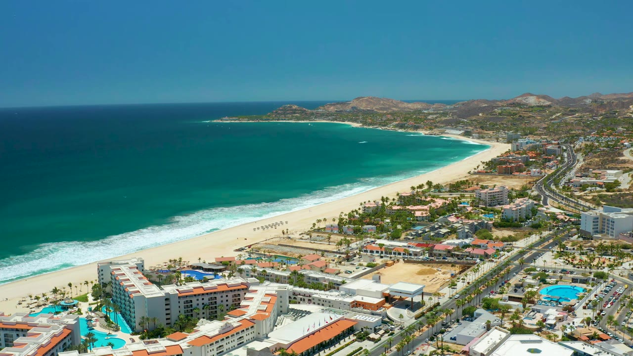 San Jose Del Cabo aerial view with the surf line slightly farther along the beach, hotels and red-roofed buildings below, blue pools near the road, low hills in the background, and no visible people.