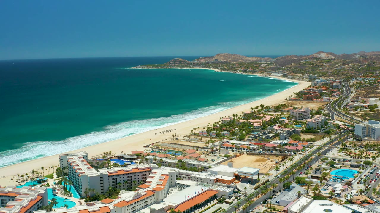 Aerial view of San Jose Del Cabo beach with deep blue and turquoise water, a bright white sand strip, beachfront buildings and pools in the lower frame, the coastal road to the right, and no visible people.