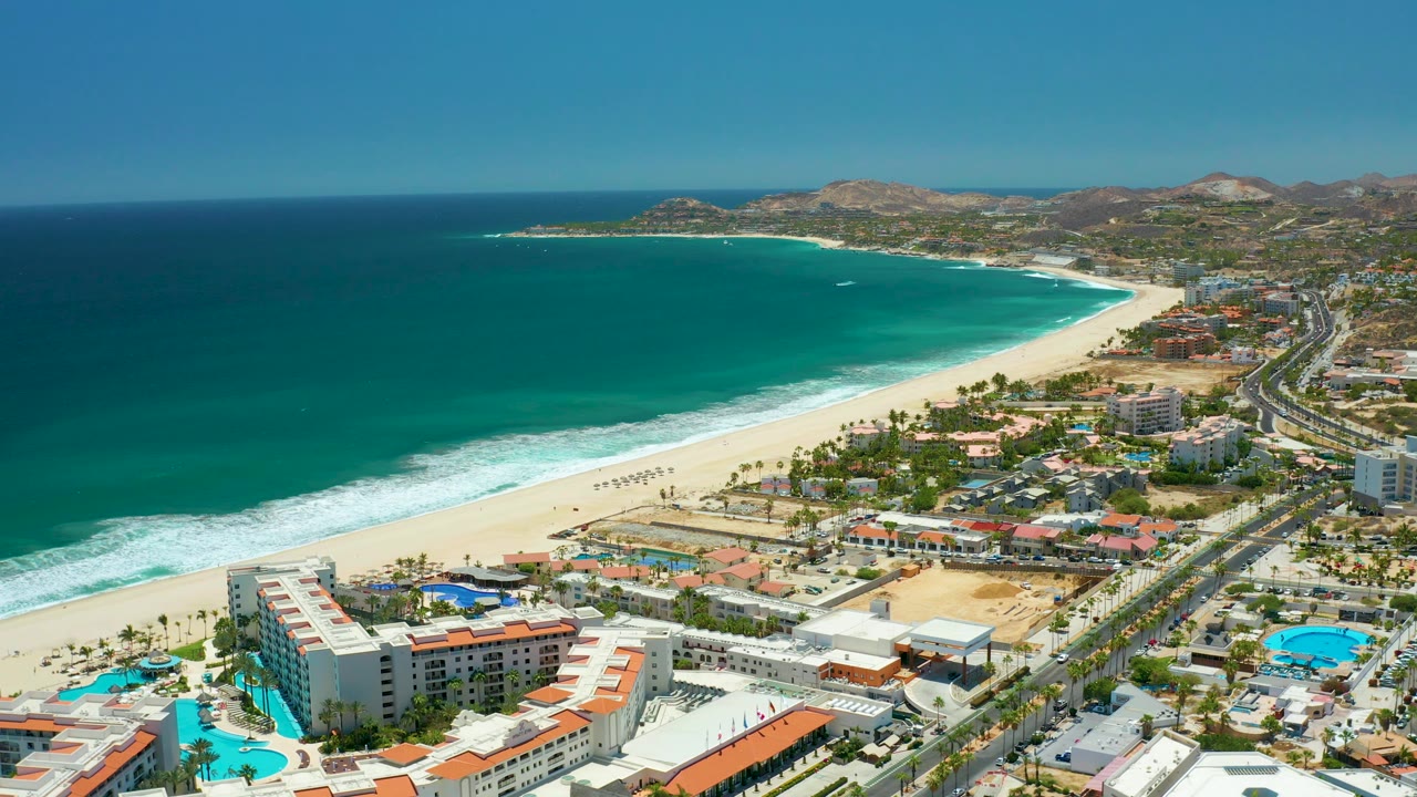Oblique drone view of a curving sandy beach, ocean waves, buildings, pools, and coastal road.