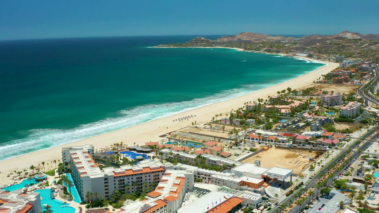 Early aerial frame in San Jose Del Cabo with turquoise water filling the left side, a curving beach through the center, resort pools and rooftops below, roads and cars to the right, hills beyond, and no visible people.