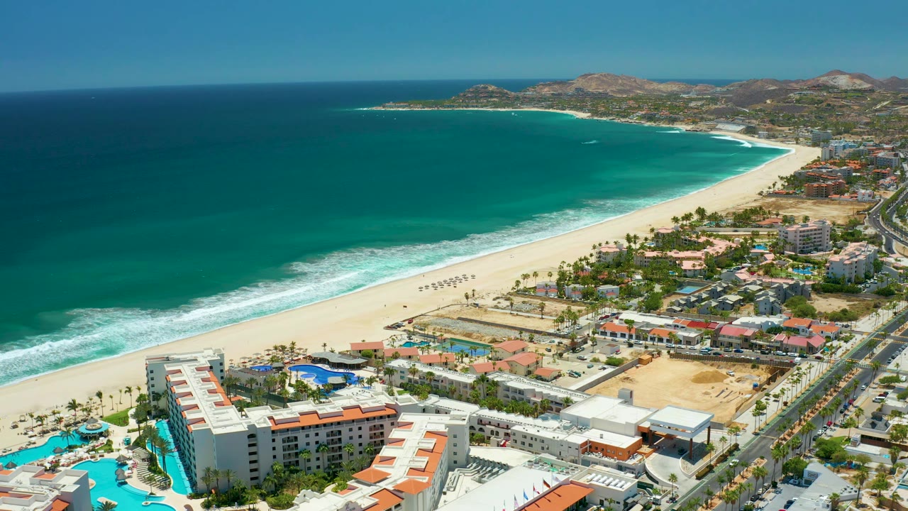 Aerial view over San Jose Del Cabo with the ocean occupying the left half, a bright beach band at center, resort buildings and blue pools below, coastal development along the right, distant hills, and no visible people.