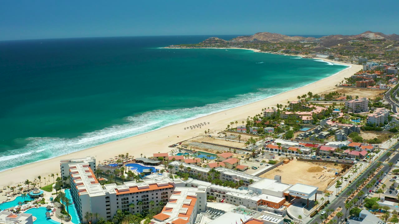 Mid-clip aerial frame in San Jose Del Cabo showing darker blue open water, a white surf edge, wide sand, resort pools and buildings at the lower edge, roadside development to the right, dry hills in back, and no visible people.