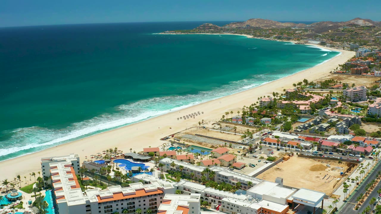 Wide San Jose Del Cabo aerial frame with the ocean covering most of the left side, white surf reaching the beach, clustered coastal buildings at lower right, and no visible people.