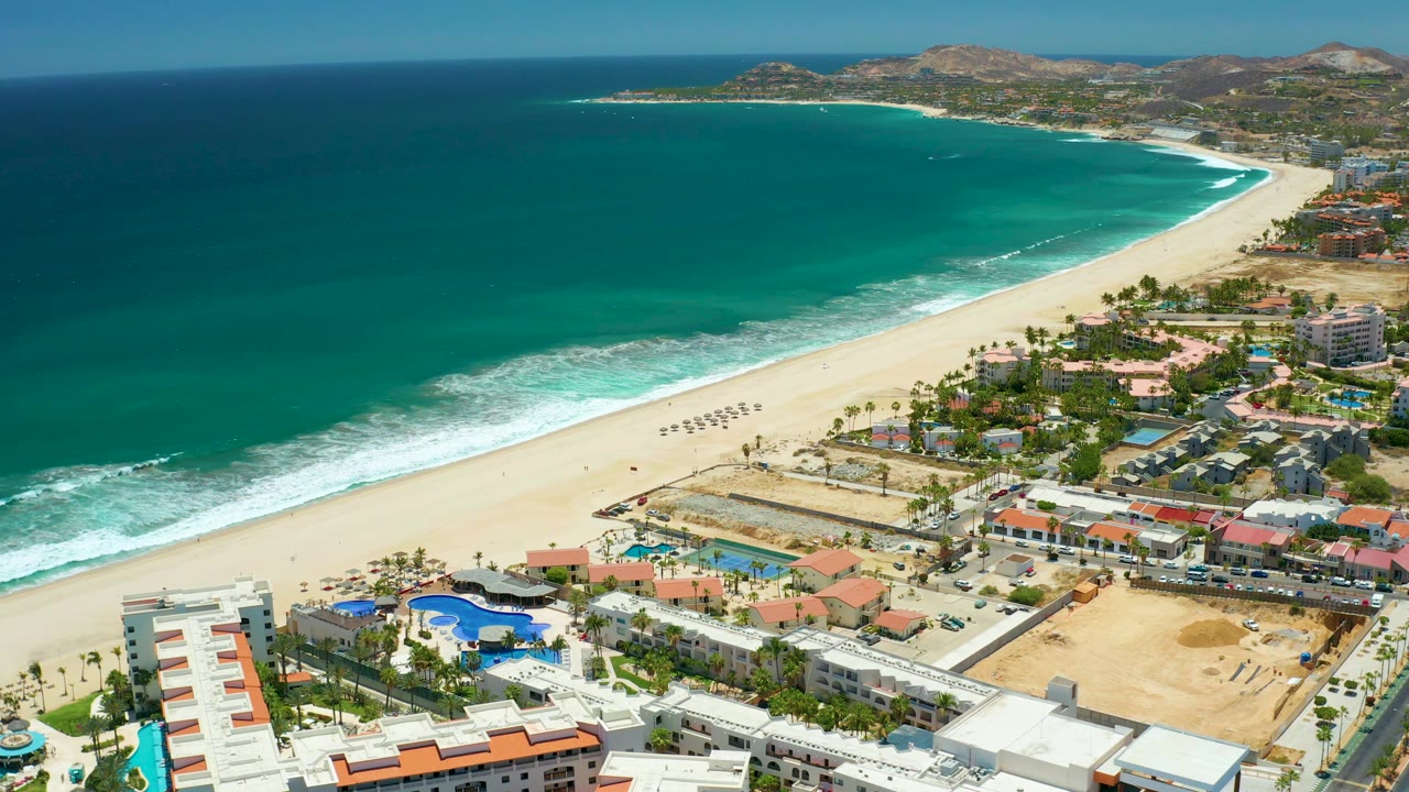 High view over San Jose Del Cabo showing ocean and surf at left, a pale sand beach through the center, resort blocks and roads in the foreground, and no visible people.