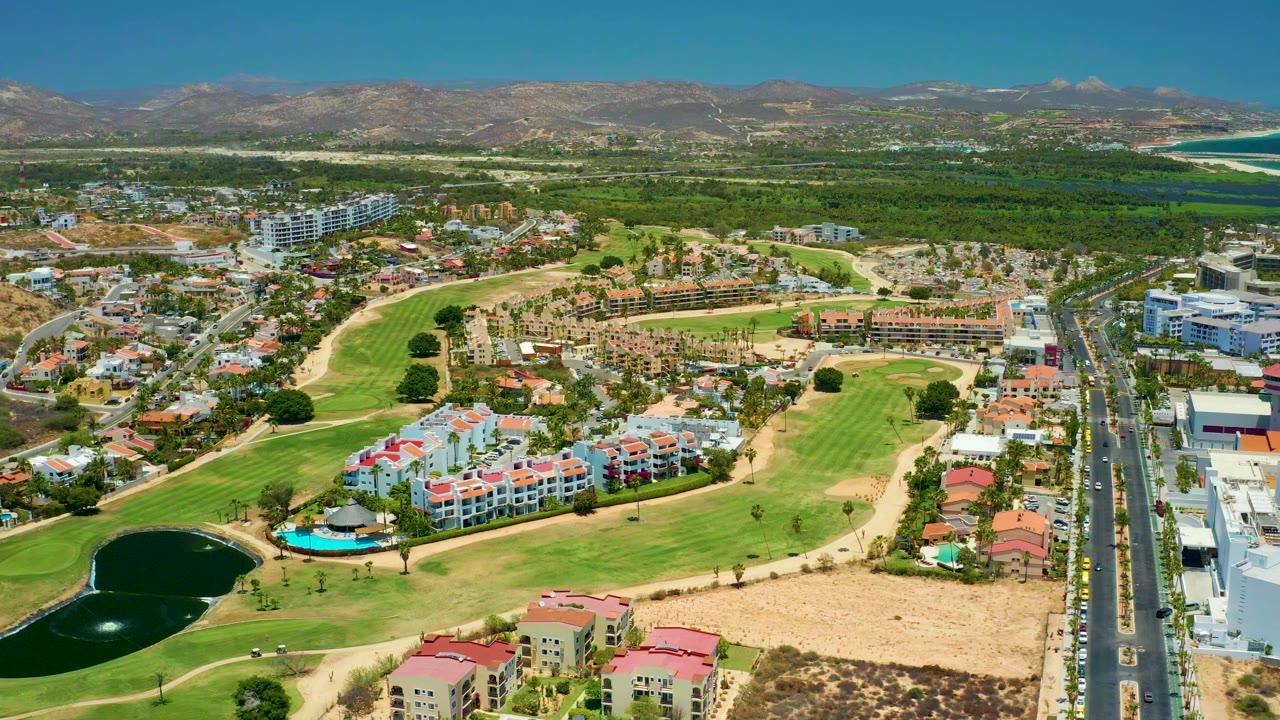 Opening aerial frame over San Jose Del Cabo with a dark pond at lower left, golf fairways, red-roofed homes, a vertical road at right, distant mountains, and no visible people.