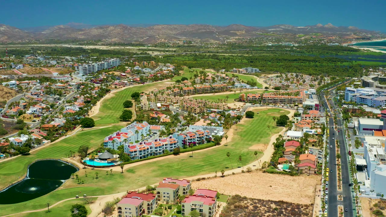 San Jose Del Cabo aerial view one second in, with the golf course and pond lower left, residential blocks across the center, road traffic on the right edge, mountains in back, and no visible people.