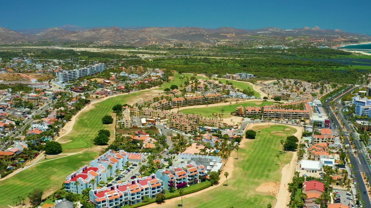 Opening aerial frame over San Jose Del Cabo showing golf fairways, white and red-roofed residential buildings, a road at right, distant mountains, coastline, and no visible people.