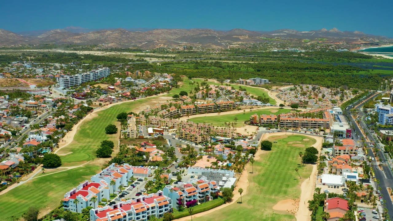 San Jose Del Cabo aerial frame showing a foreground cluster of red-roofed homes, bright golf greens, a straight road with small cars, low urban buildings, mountains, coastline, and no visible people.